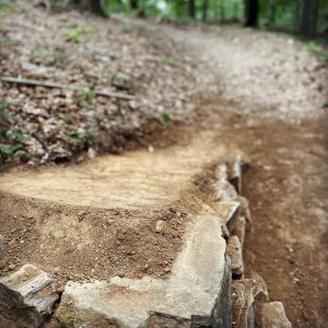 Ein Feldweg im Wald windet sich nach oben. Große Felsen säumen den Rand und dienen als Stützmauer. Der Vordergrund, ideal für Mountainbike-Fans, ist scharf und hebt die raue Textur von Erde und Stein hervor, während der Hintergrund unscharf ist.
