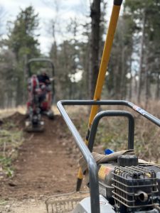Nahaufnahme einer Baumaschine mit darauf liegenden Handschuhen, positioniert auf einem Feldweg für eine neue Mountainbike-Strecke im Wald. Im Hintergrund sind eine weitere Maschine und ein Arbeiter zwischen Bäumen unter einem bewölkten Himmel zu sehen.