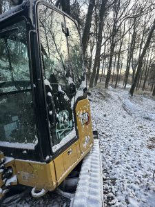 Ein gelbes Baufahrzeug mit schneebedeckter Fahrerkabine steht neben einer leicht schneebedeckten Mountainbike-Strecke im Wald. Auf der Fahrerkabinenscheibe sind Aufkleber eines Hundes und des CAT-Logos zu sehen. Im Hintergrund sind blattlose Bäume und ein verschneiter Weg zu sehen.