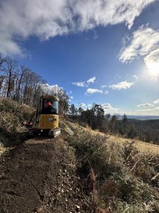 Ein kleiner gelber Bagger arbeitet auf einem Feldweg entlang eines grasbewachsenen Hügels unter einem strahlend blauen Himmel mit vereinzelten Wolken und Sonnenlicht; im Hintergrund sind Bäume und ferne Hügel zu sehen, perfekt für zukünftige Mountainbike-Abenteuer.