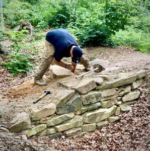 Eine Person in schwarzem Hemd und hellbrauner Hose baut eine Steinmauer entlang einer Mountainbike-Strecke in einem Waldgebiet. Sie benutzt einen Hammer und meißelt einen Stein. Werkzeuge liegen in der Nähe auf dem Boden.
