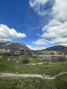 Eine malerische Landschaft mit grünen, grasbewachsenen Hügeln, vereinzelten Bäumen, einem kleinen Dorf und schneebedeckten Bergen unter einem teilweise bewölkten blauen Himmel – perfekt für eine aufregende MTB-Strecke und um den Geist der Mountainbike-Bewegung einzufangen.