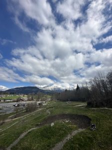 Eine Wiese mit Feldwegen und einer kleinen Grube im Vordergrund, ideal für Mountainbiker, vereinzelte Bäume rechts und schneebedeckte Berge unter einem teilweise bewölkten Himmel. Links sind einige Gebäude zu sehen.