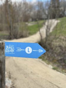 Ein blaues, pfeilförmiges Schild mit der Aufschrift „Söller Bike Saloon“ mit einem Pfeil und einem „L“ zeigt direkt neben einen gewundenen Feldweg – perfekt für jeden Mountainbike- oder MTB-Enthusiasten, der das üppige, grasbewachsene Waldgebiet erkundet.