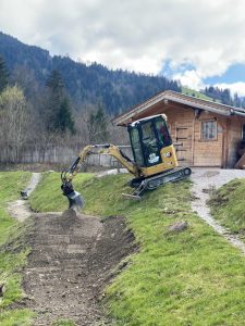 Ein kleiner gelber Bagger gräbt einen Feldweg in der Nähe eines grasbewachsenen Abhangs vor einer Holzhütte, im Hintergrund sind bewaldete Berge und ein teilweise bewölkter Himmel zu sehen – das perfekte Gelände für eine Fahrt mit dem MTB oder Mountainbike.