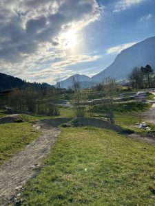 Eine sonnige Landschaft mit einem Feldweg, der sich durch grünes Gras, vereinzelte Bäume und Hügel schlängelt. Im Hintergrund erheben sich Berge unter einem teilweise bewölkten Himmel – perfekt für ein Mountainbike-Abenteuer mit durchscheinender Sonne.