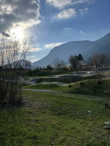 Eine Graslandschaft mit einem Pumptrack im Vordergrund, perfekt für Mountainbike-Fans, mit blattlosen Bäumen, Bergen im Hintergrund und der Sonne, die teilweise von Wolken am blauen Himmel verdeckt wird.