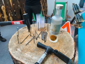 Eine Flasche klaren Schnaps, Plastikbecher, Metallnägel und zwei Hämmer liegen auf einem großen Baumstumpf entlang der Mountainbike-Strecke. Im Hintergrund sind Holzstämme, Klappstühle und ein blauer Ständer zu sehen.