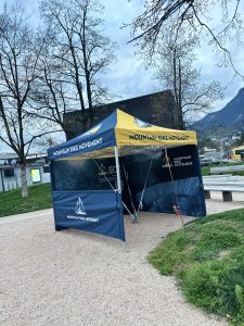 Ein blau-gelbes Pop-up-Zelt mit der Aufschrift „Mountainbikemovement“ steht neben einem Schotterweg in einem Park, im Hintergrund sind Bäume und Berge in der Ferne zu sehen.