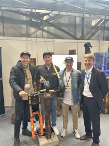 Vier Männer stehen lächelnd an einem Indoor-Eventstand. Drei tragen LOOP-Markenhüte und Freizeitkleidung, der vierte trägt einen Anzug. Sie posieren hinter einem Mountainbike-Federungsprodukt – ideal für jede MTB- oder Mountainbike-Bewegung –, das auf Holzständern ausgestellt ist.