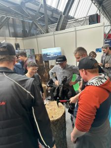 Eine Gruppe von Menschen steht drinnen um einen großen Baumstumpf herum und beobachtet, wie sich eine Person darauf vorbereitet, mit einem Hammer einen Nagel in das Holz zu schlagen. Im Hintergrund sind Messestände und Industriebalken zu sehen, die auf ein Mountainbike-Event hindeuten.