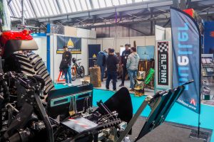 Menschen stehen und interagieren in einer Indoor-Ausstellung mit Maschinen, Holzstämmen, Bannern und Ausstellungsständen. Im Vordergrund ist ein großer Traktor neben einem Stand zu sehen, der Streckendesign und Innovationen für die Mountainbike-Bewegung präsentiert.
