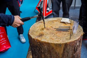 Eine Person schlägt mit einem Hammer einen Nagel in einen großen Baumstumpf. Dort liegen mehrere Nägel, ein Plastikbecher und Papiere. Andere stehen in der Nähe auf einer Art Laufstrecke, während der blaue Boden an eine Indoor-Mountainbike-Bewegungsveranstaltung erinnert.