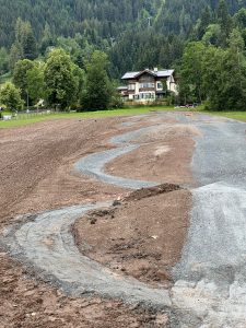 Auf einem braunen Feld aus Erde wird eine geschwungene Schotterstrecke für Mountainbikes angelegt. Im Hintergrund ist ein großes Haus zu sehen, umgeben von Bäumen und bewaldeten Hügeln.