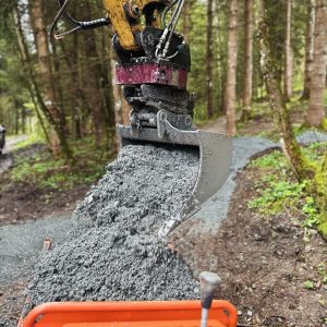 Nahaufnahme einer Baggerschaufel, die nassen Beton auf einen Wald-MTB-Trail gießt, mit Bäumen und Grün im Hintergrund. Der Weg wird in einem Waldgebiet gebaut.