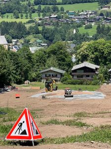 Eine Baustelle auf einer Rasenfläche mit einem dreieckigen Baustellenschild im Vordergrund, Baufahrzeuge bei der Arbeit und Häuser mit grünen Hügeln und Bäumen – ideales Gelände für eine Mountainbike-Strecke – die sich in den sonnigen Himmel erstrecken.