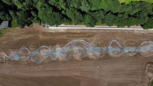 Luftaufnahme eines kurvenreichen Mountainbike-Trails im Bau auf einem gerodeten, braunen Feld, das oben von dichten grünen Bäumen gesäumt ist. Entlang der kurvigen Strecke sind Baumaterialien sichtbar.