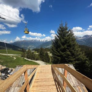 Eine Holzrampe führt hinunter in eine grasbewachsene Landschaft mit Kiefern und gewundenen Feldwegen – perfekt für Mountainbike-Abenteuer. Eine gelbe Seilbahn fährt über uns hinweg, mit schneebedeckten Bergen unter einem blauen, wolkenverhangenen Himmel.