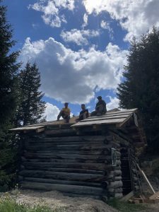 Drei Personen sitzen auf dem Dach einer rustikalen Holzhütte, umgeben von Kiefern unter einem teilweise bewölkten Himmel, mit Blick auf eine kurvenreiche Mountainbike-Strecke, während das Sonnenlicht durch die Wolken über ihnen strömt.