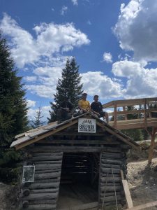 Drei Personen sitzen auf einer kleinen Holzkonstruktion mit einem Bikepark-Schild, umgeben von Bäumen und unter einem leicht bewölkten Himmel. Eine Holzrampe oder ein Mountainbike-Pfad schlängelt sich nach rechts durch die Szene.