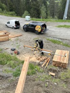 Eine Person mit Hut bedient eine Gehrungssäge auf einer Baustelle im Freien, umgeben von Stapeln aus Holz und Werkzeugen, während im Hintergrund in der Nähe eines Waldstücks zwei Fahrzeuge und ein Mountainbike auf einer Schotterstraße geparkt sind.