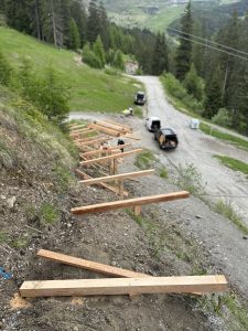 Holzbalken sind über einen abfallenden Erdhügel gelegt und scheinen das Gerüst für Treppen oder Stufen zu bilden – perfekt für einen Mountainbike-Trail. Drei Fahrzeuge parken auf einer Schotterstraße darunter, umgeben von grünen Bäumen und Graslandschaft.