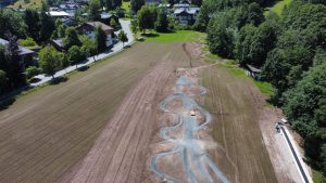 Luftaufnahme eines Feldes mit einem neu angelegten, gewundenen Schotterweg, perfekt für Mountainbike-Fans, auf der einen Seite von Bäumen und auf der anderen Seite von Häusern gesäumt, unter klarem Himmel.