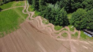 Luftaufnahme einer kurvenreichen Mountainbike-Strecke, die sich durch ein Feld und an dichten grünen Bäumen entlang schlängelt, wobei Abschnitte mit bearbeitetem Boden und Grasflecken sichtbar sind.