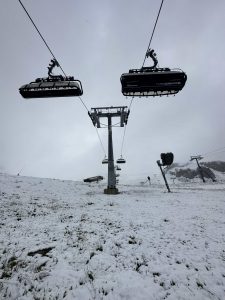 Eine verschneite Skipiste mit leeren, an Seilen hängenden Sesseln, die in einen wolkenverhangenen, grauen Himmel ragen. Der Boden ist leicht mit Neuschnee bedeckt, und die Landschaft entlang der ruhigen Piste wirkt kalt und winterlich.