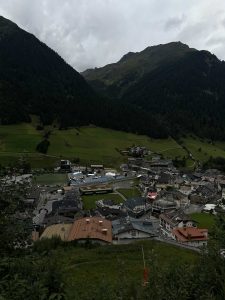 Ein kleines Dorf mit dicht beieinander liegenden Gebäuden liegt eingebettet in einem grünen Tal, umgeben von bewaldeten Hügeln und Bergen unter einem wolkigen Himmel. Ein gewundener Pfad schlängelt sich durch die Landschaft und ist perfekt für Mountainbike-Abenteuer.