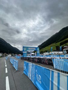 Ein Outdoor-Event-Setup mit blauen Absperrungen und Schildern markiert die Mountainbike-Strecke auf einer Straße in einer bergigen Gegend unter bewölktem Himmel. Im Hintergrund steht eine Bühne mit Mountainbikemovement-Bannern, umgeben von grünen Hügeln und Gebäuden.