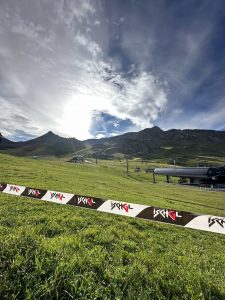 Ein grasbewachsener Berghang mit Skiliften und einer Barriere im Vordergrund, auf dem sich eine Mountainbike-Strecke unter einem teilweise bewölkten Himmel windet, während die Sonne hell über die fernen Gipfel scheint.