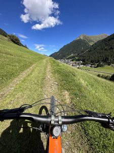 Blick aus der Perspektive eines Mountainbike-Fahrers auf einen grasbewachsenen MTB-Trail, mit sichtbarem Lenker im Vordergrund, grünen Hügeln auf beiden Seiten, einem Dorf voraus und blauem Himmel mit vereinzelten Wolken darüber.
