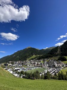 Eine malerische Aussicht auf eine kleine Stadt, eingebettet in ein grünes Tal mit bewaldeten Bergen im Hintergrund, einem klaren blauen Himmel darüber und einem großen Sportplatz mit einer Leichtathletikbahn und einem Parkplatz im Vordergrund.