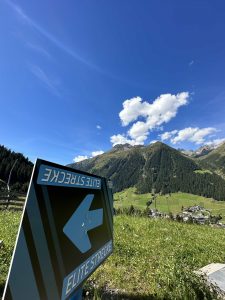 Ein blaues Schild mit der Aufschrift „ELITE STRECKE“ und einem nach links zeigenden Pfeil steht auf einer Wiese und weist Bikern den Weg zur Bergstrecke. Im Hintergrund sind unter dem strahlend blauen Himmel Berge, vereinzelte Wolken und ein Dorf zu sehen – eine Anspielung auf die wahre Mountainbike-Bewegung.