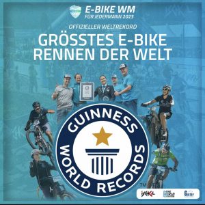 Eine Werbegrafik für die E-Bike WM 2023 zeigt Radfahrer, die mit E-Bikes auf einer spannenden Strecke fahren, Menschen, die mit einer Guinness-Weltrekord-Urkunde jubeln, und einen Text, der besagt, dass es das größte E-Bike-Rennen der Welt ist.