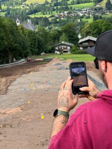 Eine Person in einem kastanienbraunen Kapuzenpullover fotografiert mit ihrem Smartphone eine Baustelle. Die Baustelle, möglicherweise der Beginn eines neuen Wanderwegs, ist mit Erde, verstreuten Steinen und bunten Fahnen bedeckt. Im Hintergrund sind grüne Hügel, Bäume und Häuser zu sehen.