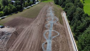 Luftaufnahme eines im Bau befindlichen Parks mit einer gewundenen, schlangenartigen Mountainbike-Strecke durch ein großes unbefestigtes Gebiet, umgeben von Bäumen und Grasflächen.