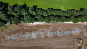 Luftaufnahme eines gewundenen Mountainbike-Trails im Bau auf einem unbefestigten Gelände, gesäumt von grünem Gras und dichten Bäumen am oberen Bildrand.