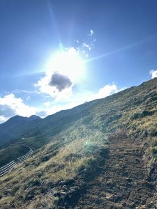 Ein sonnenbeschienener Feldweg schlängelt sich bergauf durch grasbewachsenes Gelände mit Bergen im Hintergrund, perfekt für ein Mountainbike-Abenteuer unter einem strahlend blauen Himmel mit ein paar Wolken.