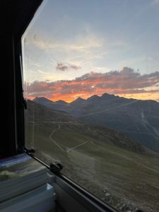 Blick aus dem Fenster auf eine Berglandschaft bei Sonnenuntergang mit orangefarbenen und rosa Wolken über schroffen Gipfeln. Ein kurvenreicher Mountainbike-Track schlängelt sich die grünen Hänge hinunter, und im Vordergrund liegen Bücher auf der Fensterbank.