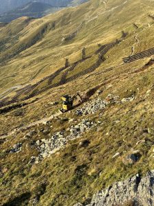 Ein kleiner gelber Bagger arbeitet an einem felsigen Berghang und formt im Sonnenlicht einen neuen Mountainbike-Trail inmitten grasbewachsener Hügel und im Zickzack verlaufender Holzzäune.
