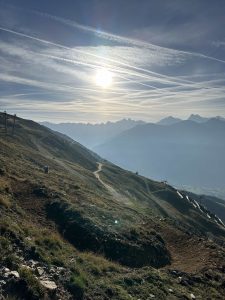 Eine malerische Berglandschaft bei Sonnenaufgang oder Sonnenuntergang mit Grashängen, gewundenen Pfaden und fernen Gipfeln, die sich vor einem von Wolken und Kondensstreifen durchzogenen Himmel abzeichnen – perfekt für jeden Mountainbike-Enthusiasten.