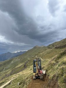 Ein kleiner Bagger gräbt unter einem bewölkten Himmel an einem grasbewachsenen Berghang, im Hintergrund sind sanfte Hügel, Holzzäune und ein gewundener Mountainbike-Pfad zu sehen.