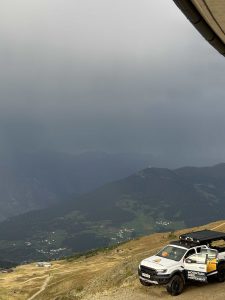 Ein weißer Truck von Mountain Management parkt unter einem bewölkten Himmel an einem Berghang und überblickt ein Tal mit verstreuten Häusern und grünen Hügeln – eine perfekte Aussicht für jeden Mountainbike-Enthusiasten, der die nächste Strecke erkundet.