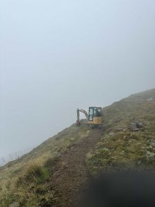 Ein kleiner gelber Bagger steht auf einem schmalen Feldweg an einem grasbewachsenen Hang, umgeben von dichtem Nebel, der den Hintergrund verdeckt und eine neblige, einsame Atmosphäre schafft – das perfekte Gelände für jedes MTB- oder Mountainbikemovement-Abenteuer.