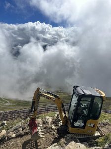 Ein gelber CAT-Minibagger steht auf felsigem Gelände hoch oben in den Bergen, teilweise umgeben von Nebel und Wolken, im Hintergrund sind grüne Hänge und ein gewundener Pfad zu sehen.
