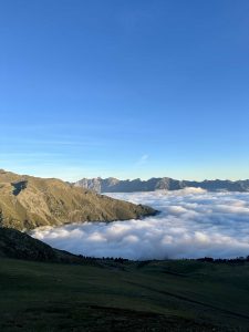 Eine malerische Aussicht auf die Berge unter einem klaren blauen Himmel, mit einer dicken Wolkenschicht in den Tälern darunter und einem gewundenen Mountainbike-Trail, der sich durch die grünen Hänge im Vordergrund schneidet.
