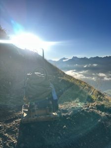 Ein Erdverdichter steht bei Sonnenaufgang auf einem Feldweg an einem Berghang, mit hellem Sonnenlicht und Berggipfeln im Hintergrund – das perfekte Gelände für jedes MTB- oder Mountainbikemovement-Abenteuer.