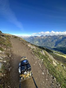 Ein unbefestigter Bergpfad, perfekt für Mountainbike-Fans, wird maschinell verdichtet, mit malerischen grünen Tälern und schroffen Gipfeln unter einem klaren blauen Himmel im Hintergrund.
